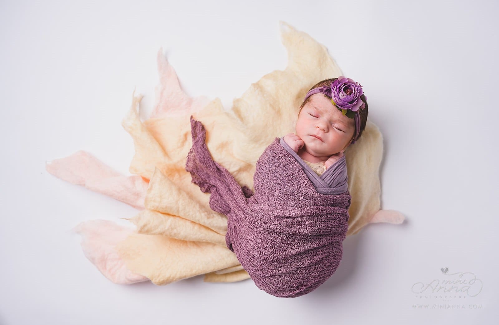 Portrait of a newborn baby girl wrapped in multiple layers of purple and ivory fabric shaped like flower.