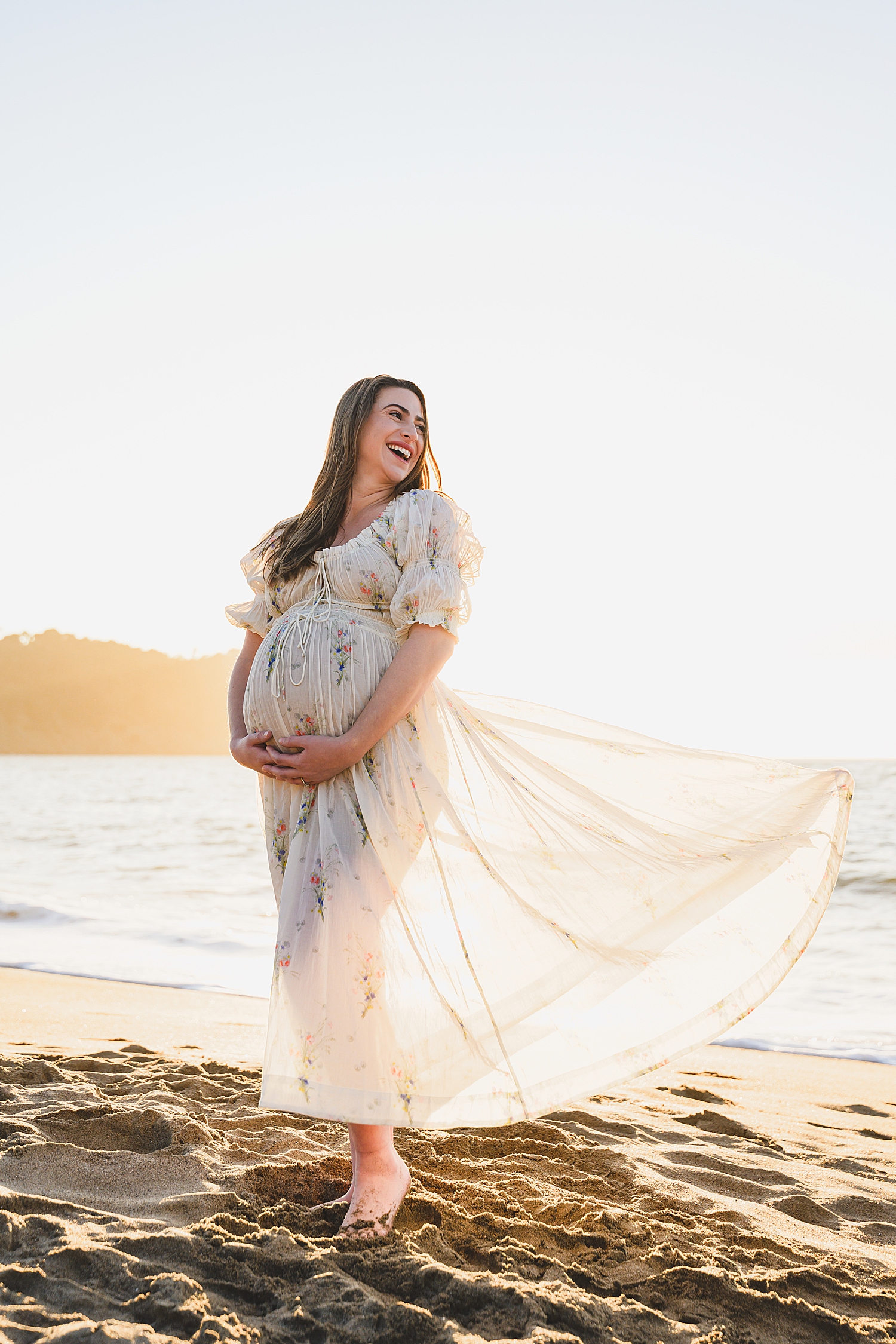 expecting mom stands on beach in white dress showing what to wear for family photos